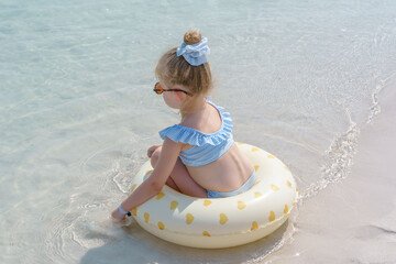 A little blonde girl with a blue gingham bow and blue-and-white striped ruffled swimsuit, sits on a cream-colored swim ring with hearts at the edge of crystal-clear turquoise water 