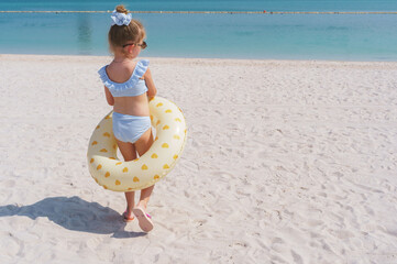 Little girl with swim ring from behind walks across a white sandy beach toward the turquoise sea or ocean 