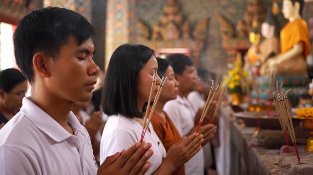 A group of devotees hold incense while praying in a temple where devotees face a golden Buddha. The devotees' hands with incense contribute to the spiritual atmosphere in the templ
