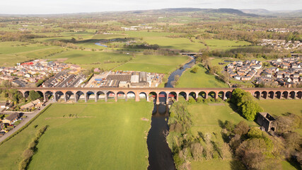 Aerial view of a colossal red brick viaduct slicing through vibrant green fields and reflecting in the dark ribbon of river below, Whalley, England, United Kingdom.