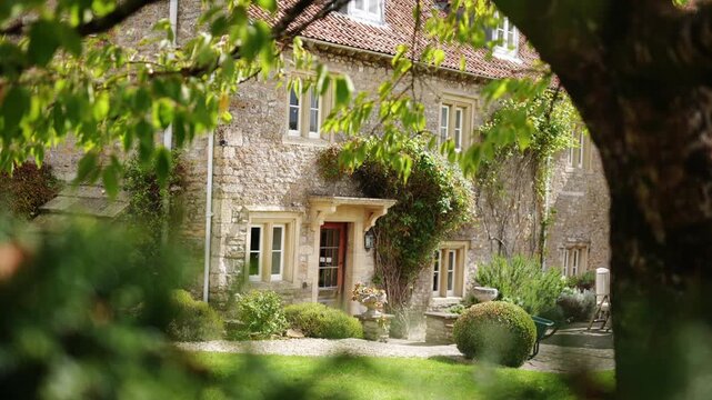 A large stone house with a large tree in front of it. The house has a lot of windows and a large front door. The tree is full of leaves and branches, and it is casting a shadow on the ground