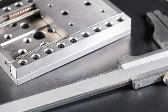 Close up of a precision linear motion guide rail with metal screws and a caliper on a black background.