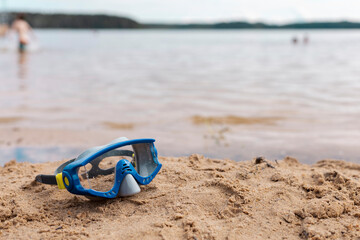 Bright blue snorkeling gear lies on soft sand by the water. People can be seen enjoying the water in the background under a clear sky, creating a peaceful scene