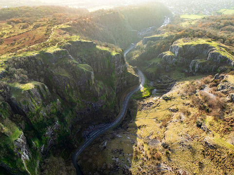 Aerial view of sunlight casting long shadows over the rugged cliffs and winding road cutting through Cheddar Gorge's dramatic landscape, Cheddar, England, United Kingdom.