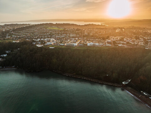 Aerial view of the sun casting a golden hue over the vast forest and the adjacent coastal town, nestled by the serene sea, Babbacombe, England, United Kingdom.