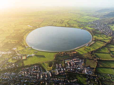 Aerial view of a serene reservoir reflecting the sky, surrounded by vibrant green fields and nestled near a charming village, Cheddar, England, United Kingdom.