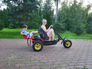 A woman and a young boy enjoy a ride together on a pedal go kart in a park surrounded by green trees and grass. A child aged 4 years (four years old)