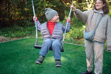 Woman helping a little kid on a swing. Happy caucasian boy having fun on a playground. Family leisure activity outdoors. A child aged 4 years (four years old)