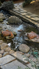 A serene Japanese garden featuring a tranquil lake surrounded by lush greenery and colorful autumn leaves. Sculptures are placed throughout the landscape.