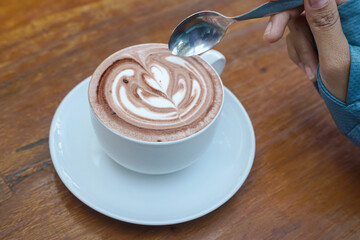 Cappuccino coffee with heart-shaped latte art on a wooden table, a close-up photo of a cappuccino drink in a white cup with a spoon on a wood background.