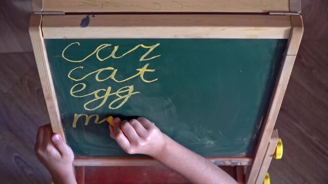 Top view closeup of a 9-year-old child writing cat, car, egg, milk, dog, bread on a chalkboard with chalk at home.