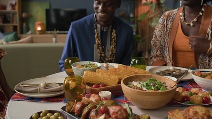 Medium shot of young Black woman placing corn on colorful Kwanzaa table as she joining family to celebrate with traditional food, unity, and cultural heritage in warm, festive home setting - Powered by Adobe