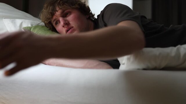 Young man lies tired on a bed with his head on a pillow looking away with exhausted expression showing fatigue stress and burnout symbolizing unhealthy lifestyle and lack of motivation