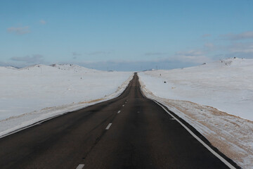 Empty highway stretching into the distance through snow-covered hills under a clear blue sky, symbolizing travel and solitude