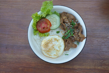A vibrant bowl of rice topped with grilled beef, a fried egg, and colorful vegetables, served in a rustic dish