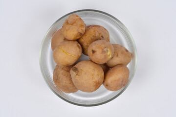 A glass bowl is filled with whole boiled potatoes with their light brown skins on, centered against a solid white background.