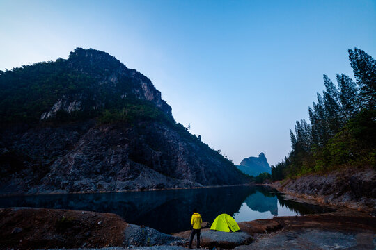A serene camping scene with a bright green tent and a traveler by a calm mountain lake at dawn, surrounded by rugged cliffs and pine trees reflecting on the water.