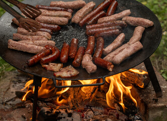 A large, round, flat metal grill, discada or plow disc grill, being used over an open flame. Sausages, pork and beef steaks, and kebabs are cooked on it. A little greenery is visible in the background