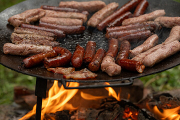 A large, round, flat metal grill, discada or plow disc grill, being used over an open flame. Sausages, pork and beef steaks, and kebabs are cooked on it. A little greenery is visible in the background