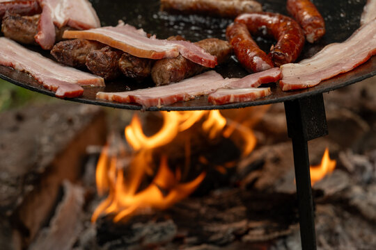 A large, round, flat metal grill, discada or plow disc grill, being used over an open flame. Sausages, pork and beef steaks, and kebabs are cooked on it. A little greenery is visible in the background