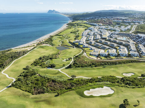 Aerial view of verdant golf courses meet the turquoise sea under the gaze of distant mountains, La Alcaidesa, Andalusia, Spain.