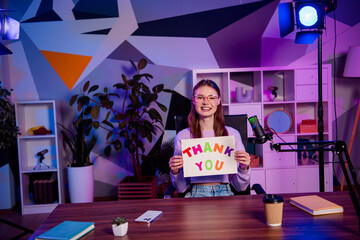 Young female blogger holding thank you sign in colorful studio with creative decor and professional podcasting setup