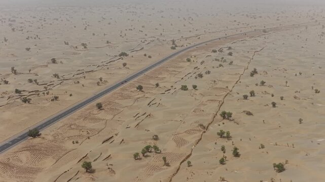 Aerial view of a dark asphalt road crossing the vast, undulating expanse of the sandy Taklamakan Desert, with sparse vegetation, Taklamakan Desert, China.