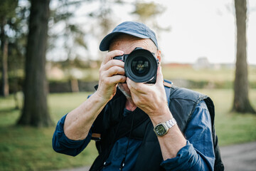 An elderly 66-year-old man with a beard wearing a cap, photographing with a camera outside in the late afternoon.