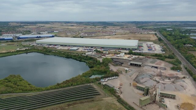 Aerial view of a vast warehouse next to a dark lake, solar panels, and a gravel pit under an overcast sky, Bedford, England, United Kingdom.