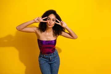 Young stylish woman posing in bright yellow studio wearing purple sequin top making peace sign and playful kiss for fashion lifestyle image