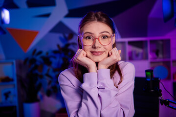 Young woman in neon-lit home office holding thoughtful pose in front of microphone setup for vlogging or podcasting