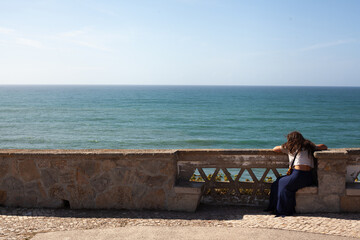 Young woman sitting alone on a stone bench, overlooking the calm blue ocean on a clear sunny day.