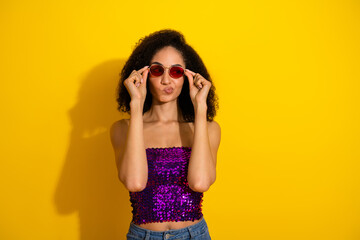 Playful glamorous young woman in purple sequin top posing with red sunglass against bright yellow background
