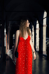 Obraz premium A woman in a vibrant red sundress with a small white floral pattern walks through a dark, arched corridor, bathed in a ray of sunlight.
