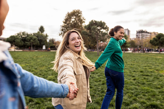 Cheerful multiracial female friends holding hands, leading and following, enjoying time together in a city park