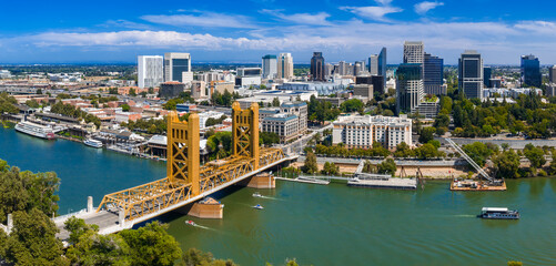 Aerial view of Sacramento, California, showing the golden Tower Bridge over the Sacramento River, downtown buildings, and the State Capitol surrounded by trees. © Aerial Film Studio