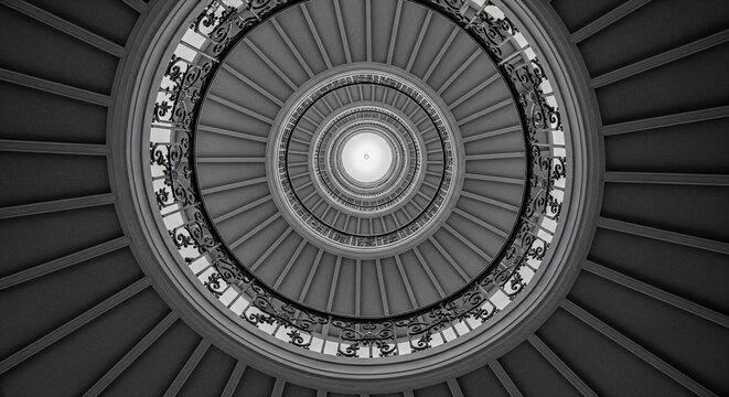 Architectural detail of a spiral staircase shot from the bottom looking up, emphasizing the elegant geometric pattern. Monochrome conversion.