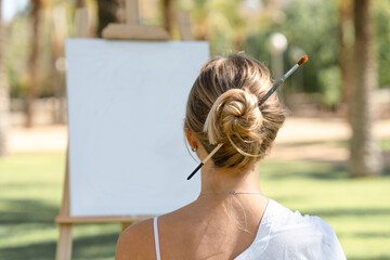 Young woman sitting outdoors with a paintbrush tucked into her hair bun, facing a blank canvas on...