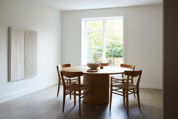 Fototapeta premium Bright dining room with round wooden table, matching chairs, and large window overlooking greenery. 