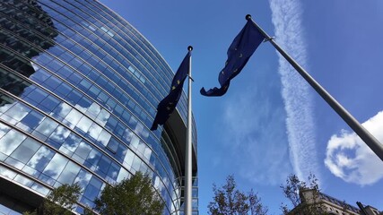 EU flags wave under a blue sky beside the curved glass facade of the Lex Building in Brussels’ European Quarter. - Powered by Adobe