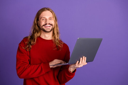 Young handsome male with long blonde hair in a red sweater smiles while holding a laptop against a purple background