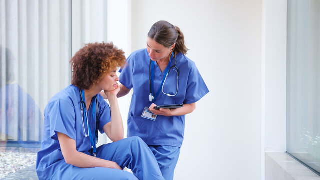 Female Doctor Wearing Scrubs Concerned About Stressed Colleague In Modern Hospital