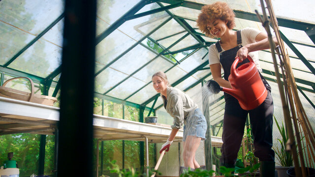 Two Female Friends Or Same Sex Couple Working In Greenhouse At Home Sweeping And Watering Plants - Powered by Adobe