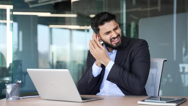 Unhealthy businessman suffers from ear pain while sitting in office. Sick male in formal suit touches his ear with his fingers. He has furuncle, shingles, eczema, ear diseases or otitis media. Earache