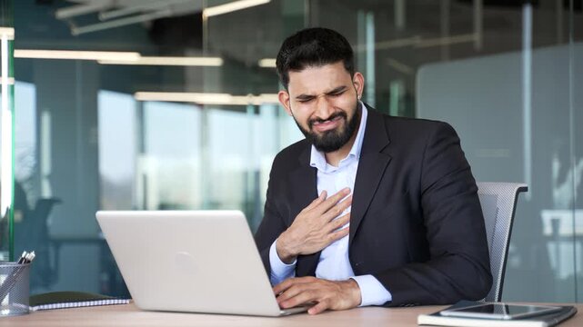 Unwell businessman suffers from heartburn sitting at workplace in business office. Unhealthy man with discomfort holding his chest with his hand, experiencing pain and grimacing, has a health issue