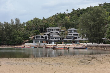 Traditional Thai fishing boats anchored near modern villas and resorts on the tropical coastline of Koh Phangan island, Thailand.