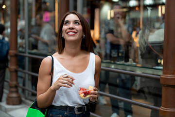 Smiling young woman holding fruit cup and looking upwards outside crowded marketplace