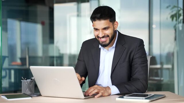 Satisfied happy businessman received great news on laptop while sitting at workplace in modern business office. Smiling joyful male worker in formal suit reads a positive good message on the computer