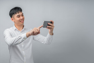 A handsome young Asian man smiles happily while using a social media or gaming app on a modern smartphone. This concept is ideal for technology, connectivity, lifestyle, and leisure.