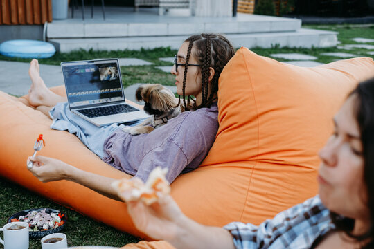Young woman on an orange bean bag holds slice of pizza and smiles at her friend. Girl works on a laptop with small dog in her lap